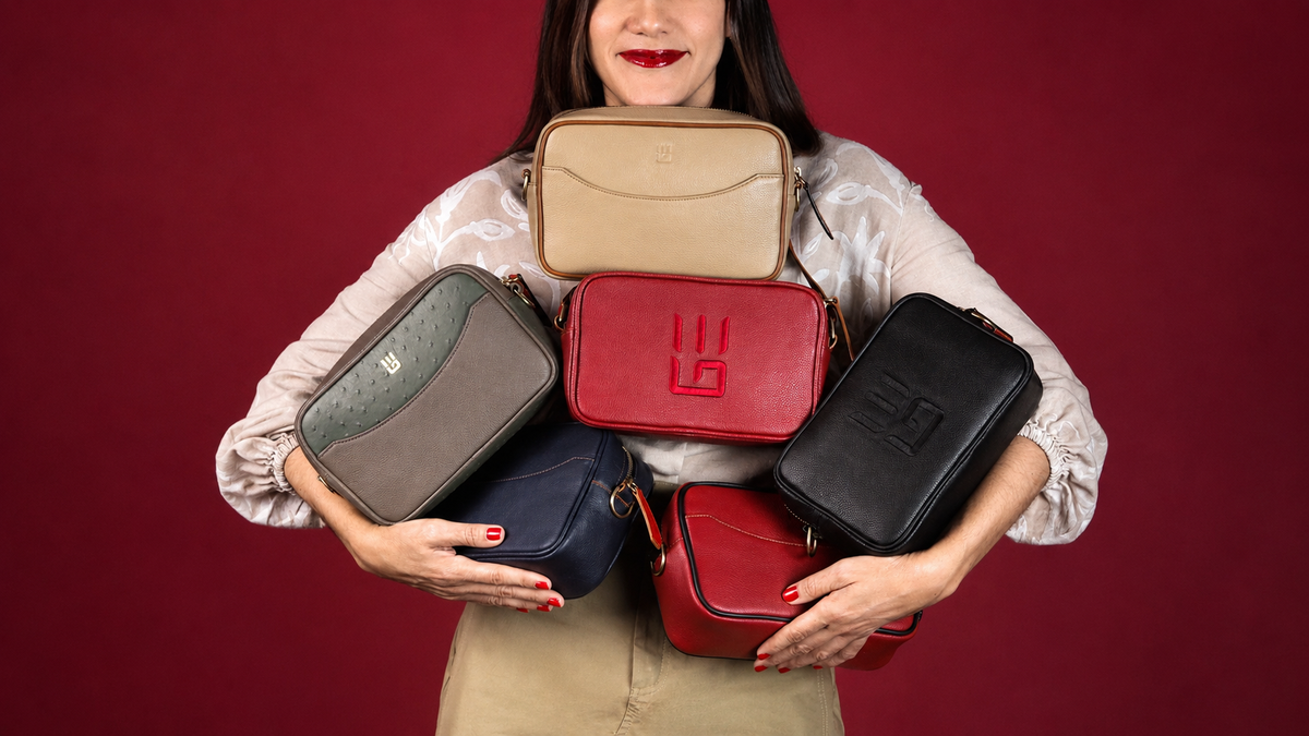A woman holds six small crossbody bags in different colors against a solid dark red background. The bags include beige, gray, navy, red, and black, with one red bag featuring an embossed monogram logo.